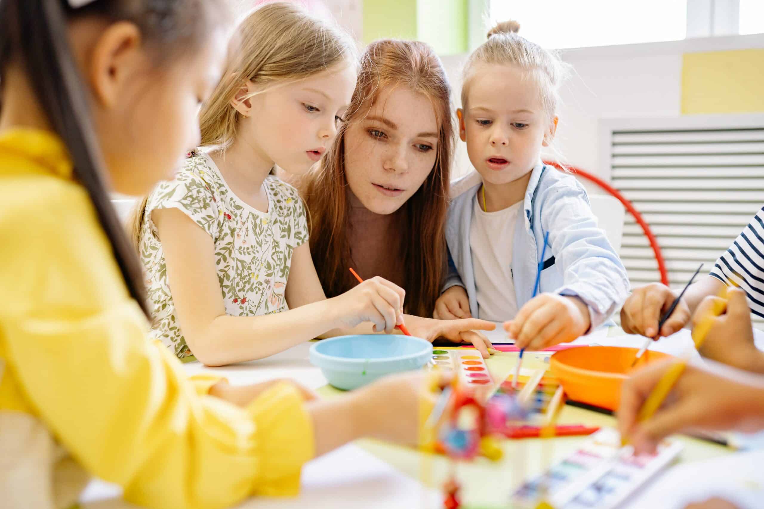 Junge Erziehering mit einer Gruppe von Kindern beim Malen am Tisch im Kindergarten.