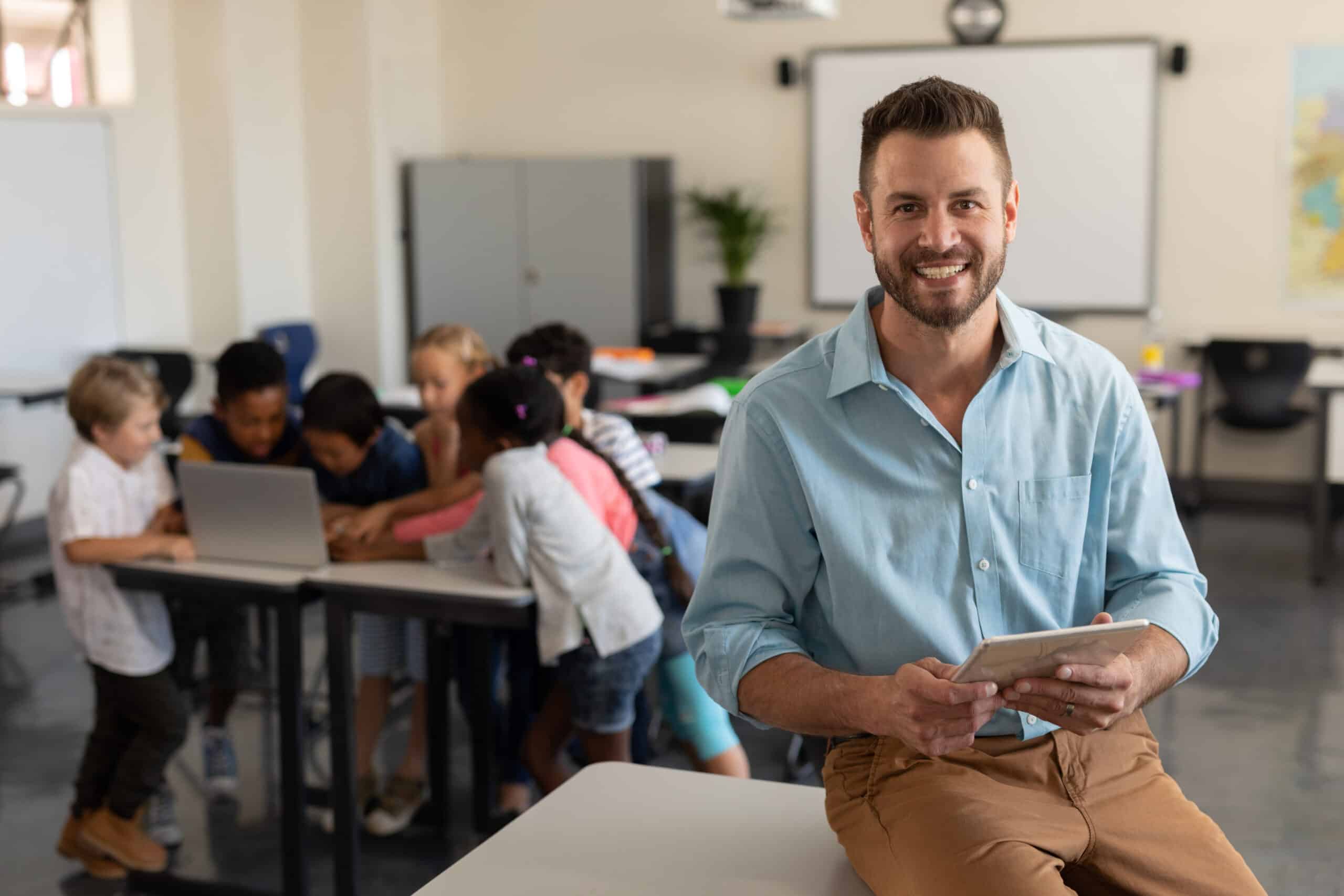Junger Erzieher mit Tablet in der Hand in einer Grundschulkasse mit Kindern am Computer im Hintergrund.