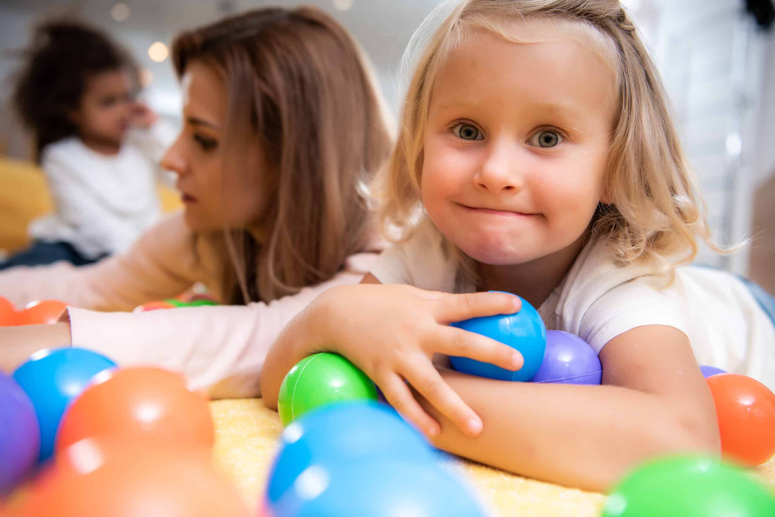 Lächelndes Mädchen mit bunten Bällen mit Erzieherin im Kindergarten.
